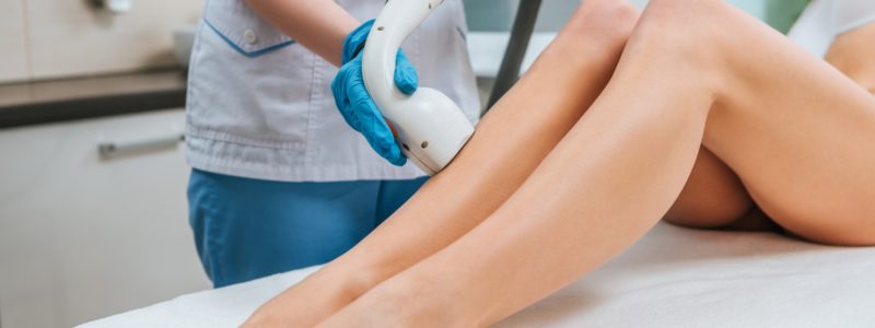 Partial view of cosmetologist in rubber gloves doing laser hair removal procedure on legs